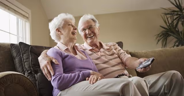 An elderly couple sitting closely together on a brown couch in a living room, smiling and laughing. The man is holding a TV remote control, and there is a window with blinds and a plant in the background.