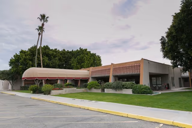 Exterior view of a single-story building with a covered entrance and a tiled roof section, surrounded by green grass, trees, and a parking lot in front under a cloudy sky.