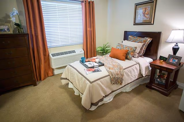 Neatly arranged single bedroom featuring a twin bed with decorative pillows, a nightstand with a lamp, a dresser, and a window with orange curtains.