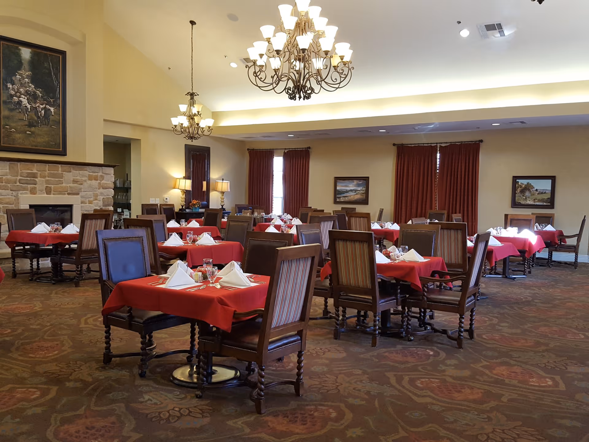 A dining room with multiple tables covered in red tablecloths, each set with white folded napkins, glasses, and silverware. The room features ornate chandeliers, a stone fireplace, framed paintings on the walls, and large windows with red curtains.