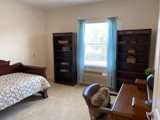 A bedroom with a single bed on the left side covered with a patterned bedspread. There are two dark wooden bookshelves on either side of a window with light blue curtains. Below the window is an air conditioning unit. On the right side of the room is a wooden desk with a chair that has a furry cushion. The room has beige walls and carpeted floor.