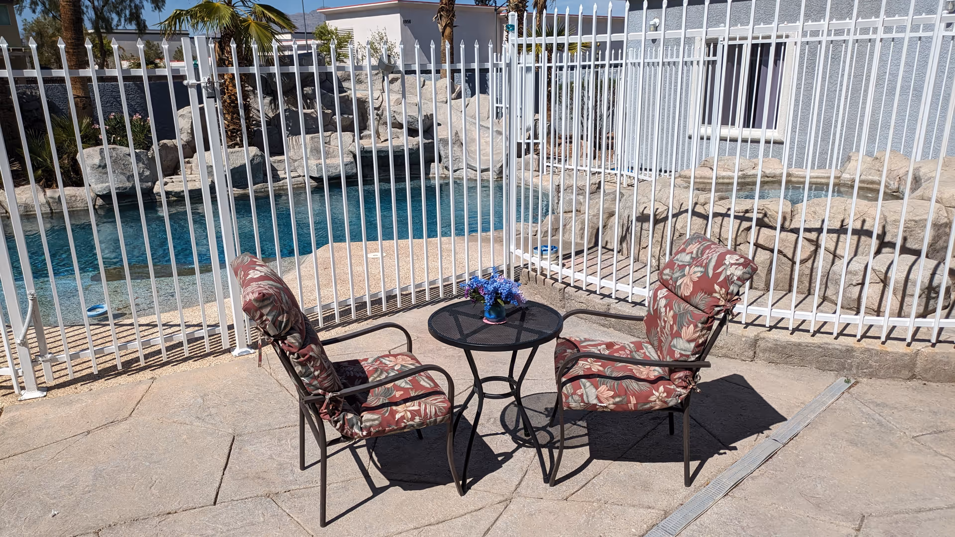 Outdoor patio area with two cushioned chairs and a small round table with a vase of flowers, situated next to a fenced swimming pool and rock waterfall feature at Mother's Touch Senior Home.