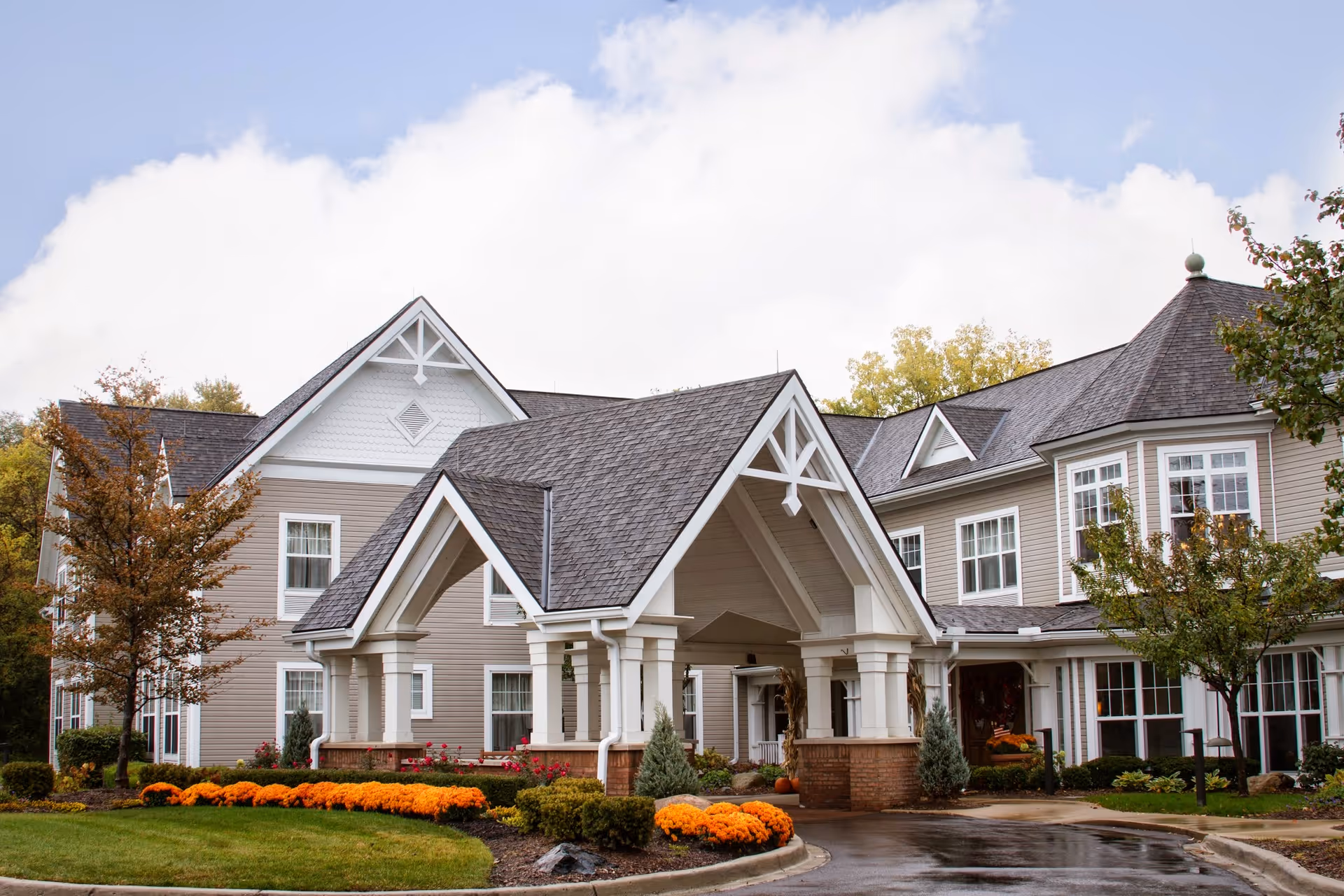Exterior view of a senior living facility building with a covered entrance, beige siding, white trim, and a landscaped garden with orange flowers and trees under a partly cloudy sky.