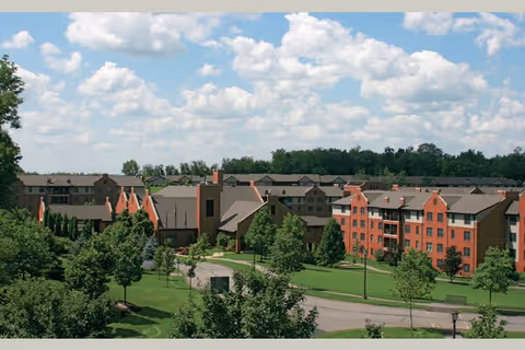 View of Masonic Village at Sewickley showing multiple residential buildings surrounded by green lawns, trees, and a clear blue sky with scattered clouds.