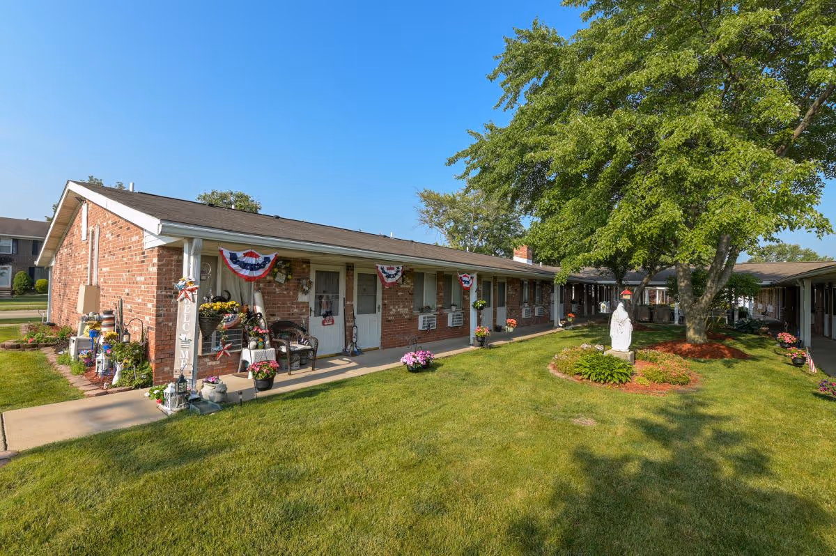 Exterior view of a single-story brick building at Maryhaven facility with a well-maintained lawn, a large tree, and a statue in the garden area. The building has several doors and windows, decorated with patriotic bunting and various potted plants and flowers along the walkway.