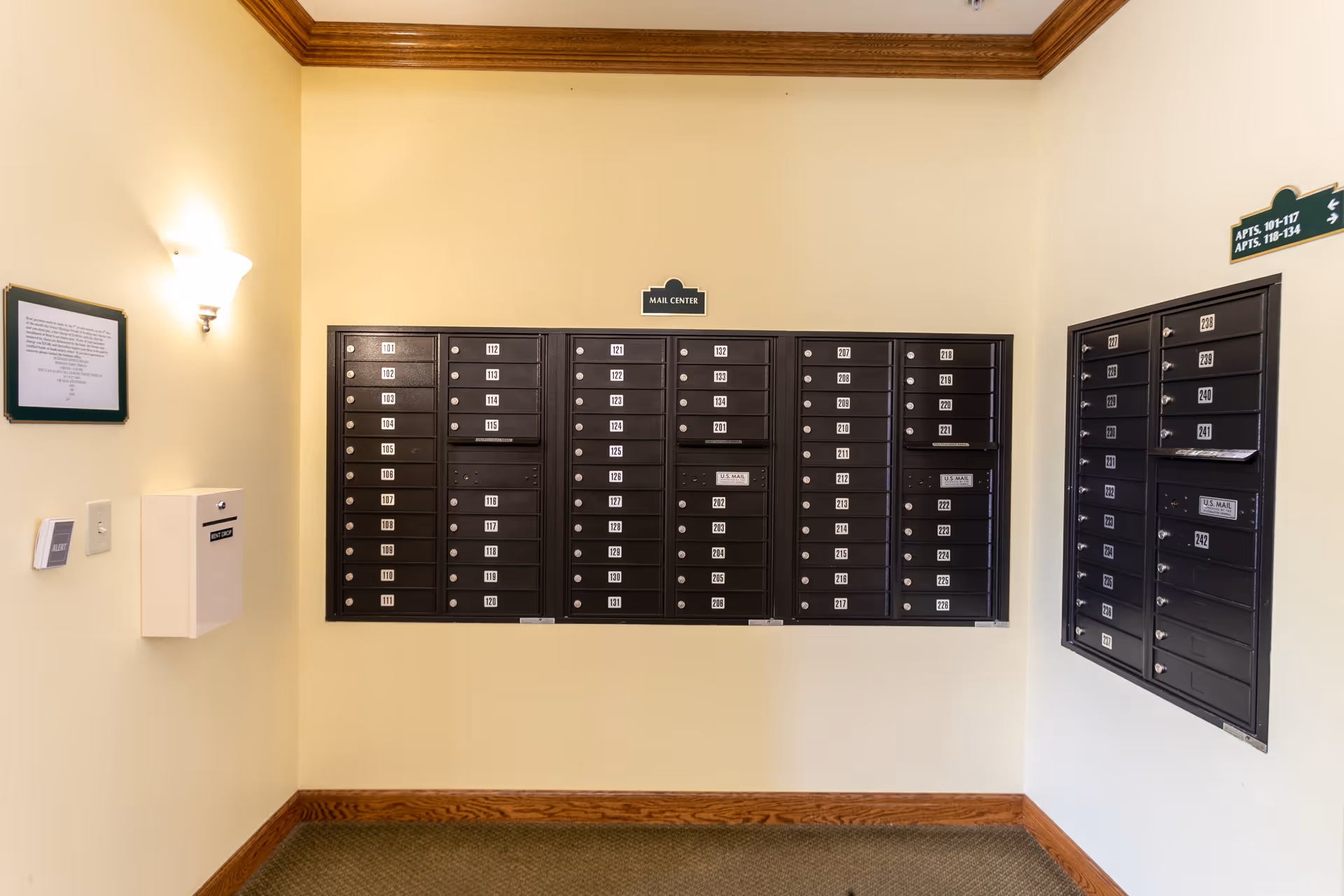 Interior view of a mail center in a senior living facility with multiple black mailboxes mounted on beige walls. The mailboxes are labeled with apartment numbers. There is a wall-mounted light fixture on the left side and a small white box below it. A sign above the mailboxes reads 'MAIL CENTER'.