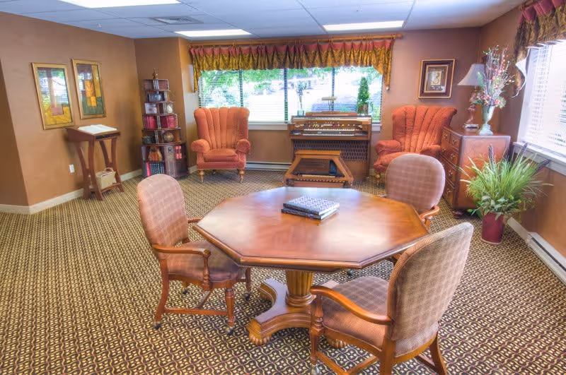 A cozy interior room with a wooden octagonal table surrounded by four upholstered chairs. In the background, there are two orange armchairs, a wooden organ, a small bookshelf, and a side table with a lamp and decorative flowers. The room has patterned carpet, brown walls, and large windows with valances letting in natural light.