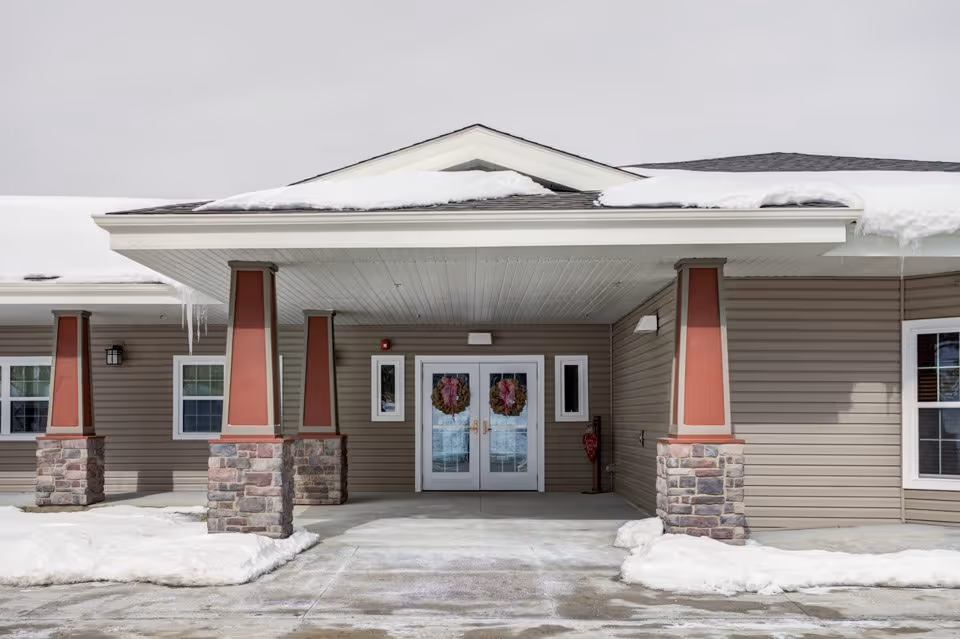 Front entrance of a single-story building with a covered porch supported by stone and red pillars, snow on the ground and roof, and double glass doors decorated with wreaths.