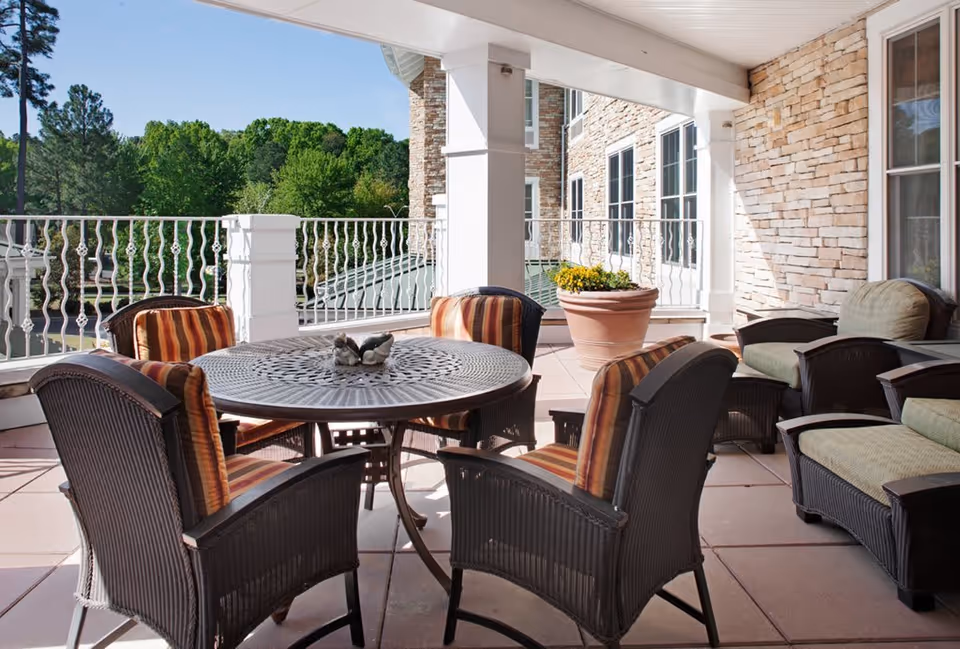 Outdoor covered patio area with a round metal table surrounded by six cushioned wicker chairs, two cushioned wicker armchairs, and a large potted plant. The patio overlooks green trees and has a stone exterior wall with windows.