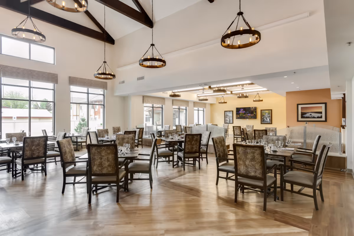A spacious and well-lit dining room with multiple tables and chairs arranged neatly. The room features large windows allowing natural light to fill the space, wooden flooring, and decorative hanging light fixtures. The walls are adorned with framed artwork, and there are semi-private booth seating areas along one side.