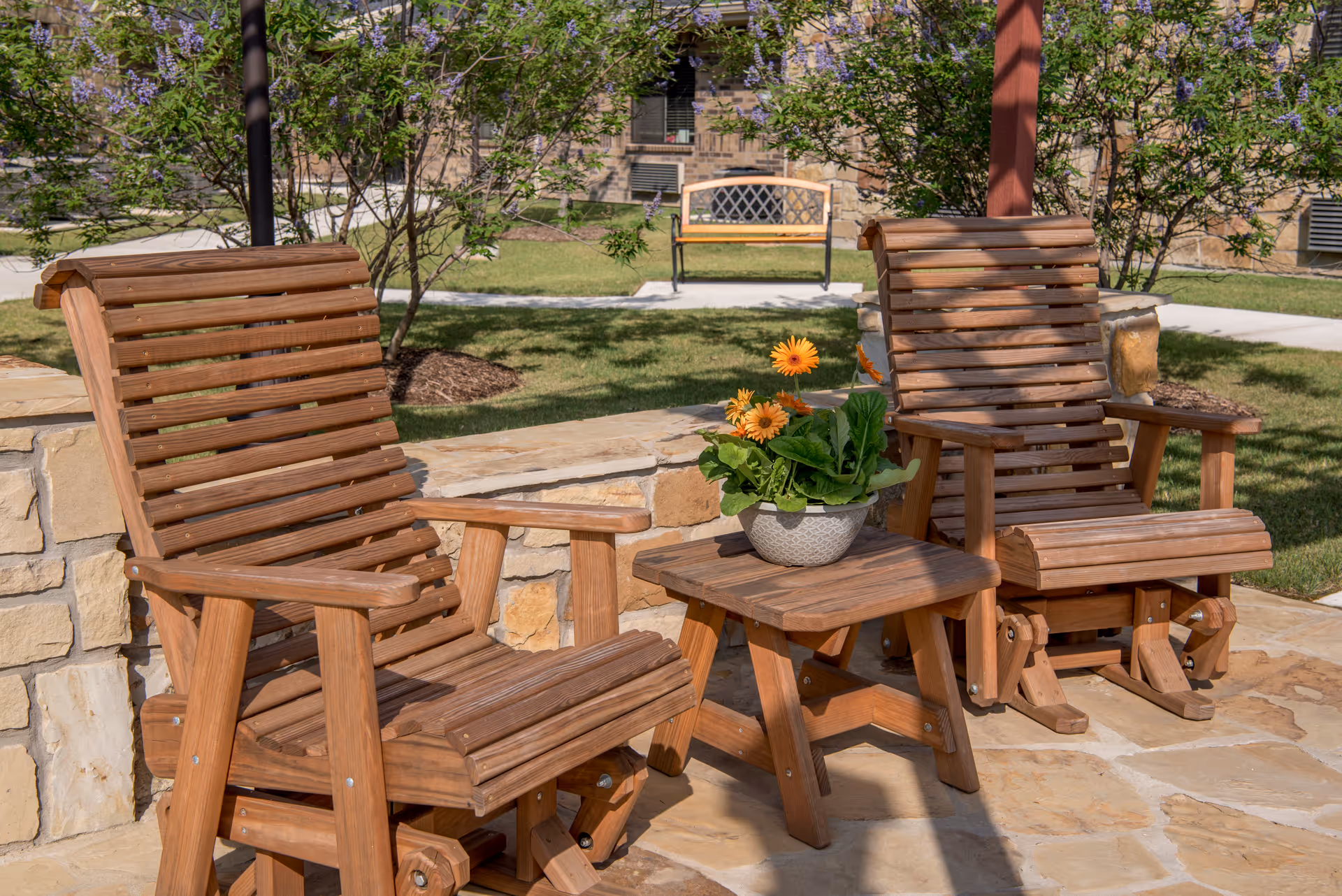 Two wooden rocking chairs with armrests and a small wooden table between them holding a pot of orange flowers, set on a stone patio with green grass, trees, and a bench in the background.