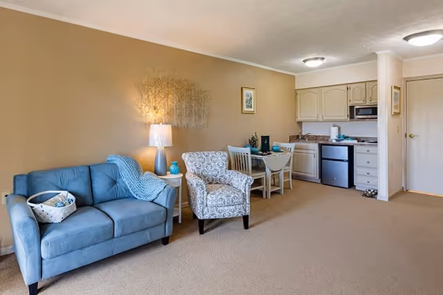 A cozy living area in a senior living facility featuring a blue loveseat with a basket on it, a patterned armchair, a small dining table with two chairs, and a compact kitchen with cabinets, a microwave, and a mini fridge. The walls are painted beige and decorated with a metal wall art and framed pictures. The room is carpeted and well-lit with ceiling lights and a table lamp.