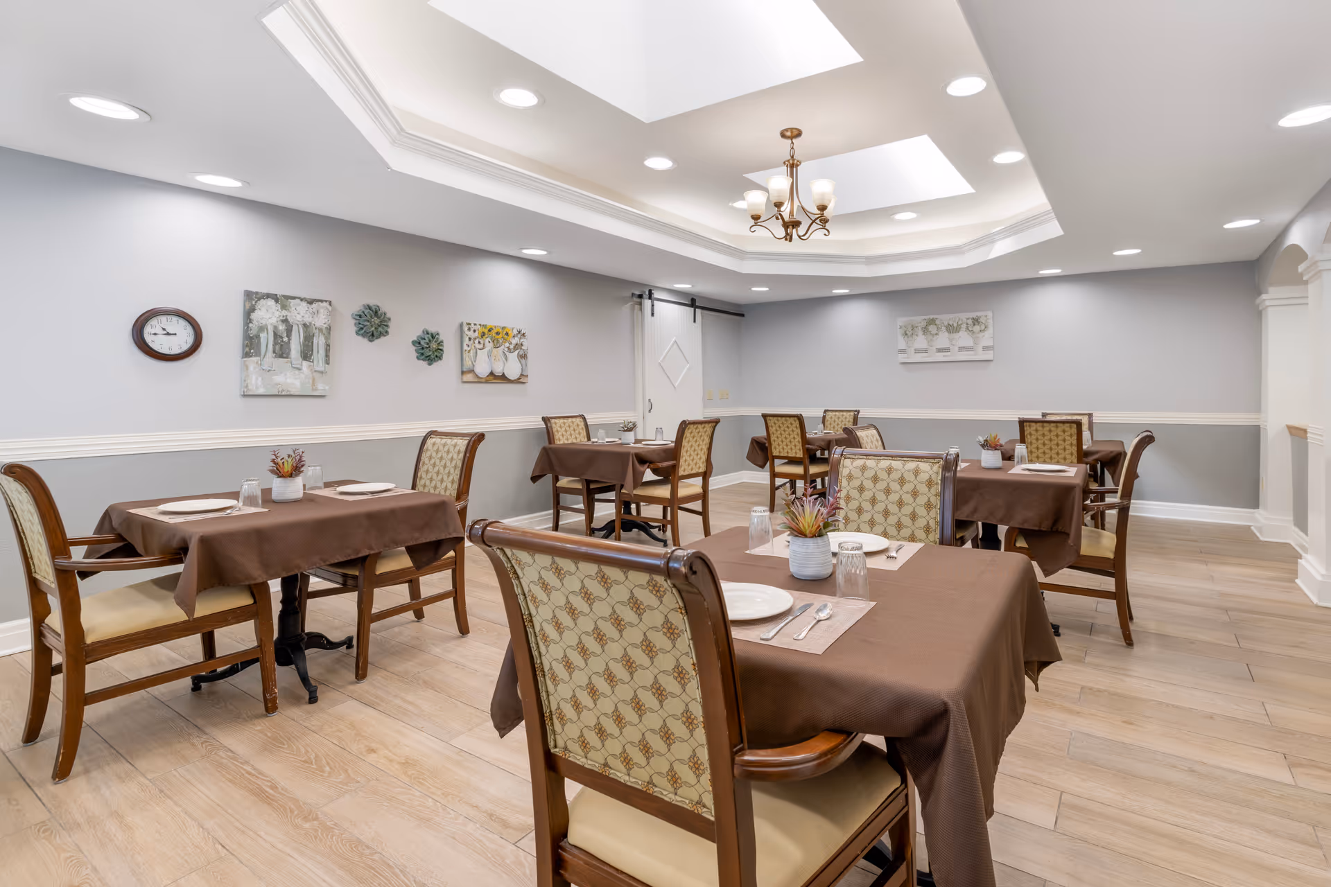 Dining room with several tables covered in brown tablecloths, upholstered wooden chairs, place settings, and recessed ceiling lighting.