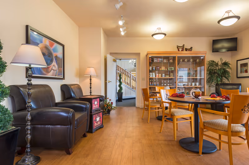 A cozy senior living common area with wooden flooring, two black leather armchairs with a floor lamp beside them, a round wooden table with four chairs set with red napkins and decorative mugs, a glass cabinet displaying various items, a potted plant, and a wall-mounted TV in the corner.