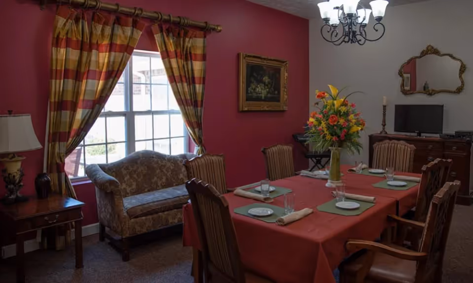 A formal dining room with a red tablecloth-covered table set for six, a floral centerpiece, chandelier, and a sofa by a window with striped curtains.