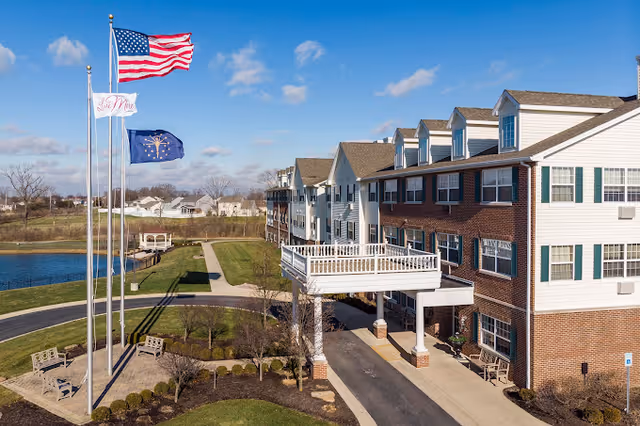 Exterior view of a multi-story senior living facility building with a covered entrance and a balcony. Three flagpoles display the American flag, a white flag with red text, and a blue flag with a yellow emblem. The surrounding area includes a paved driveway, benches, landscaped greenery, and a small pond under a partly cloudy sky.