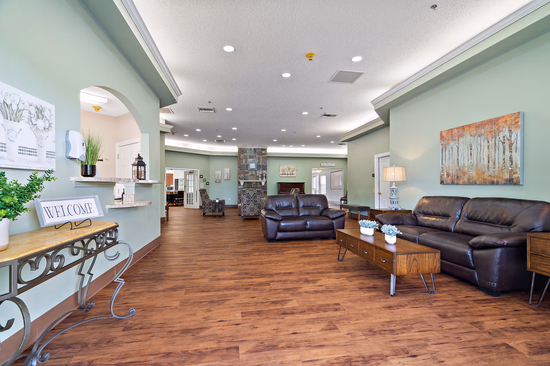A spacious living room area with wooden flooring, light green walls, and recessed ceiling lights. The room features two dark brown leather sofas, a wooden coffee table with small potted plants, a side table with a lamp, and a decorative painting of trees on the wall. There is a fireplace in the background and a console table with a 'WELCOME' sign and plants near an open window-like counter.
