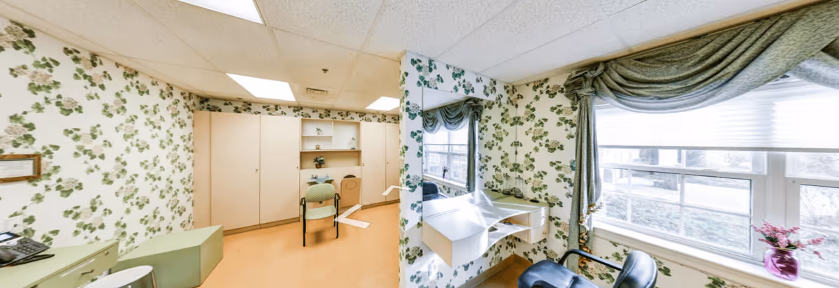 A bright room with floral wallpaper featuring green leaves and flowers. The room has a light wood floor and built-in cabinets along one wall. There is a green chair in front of the cabinets and a small desk area with a mirror on the adjacent wall. A window with sheer white blinds and green drapes lets in natural light, and a small purple vase with flowers sits on the windowsill.