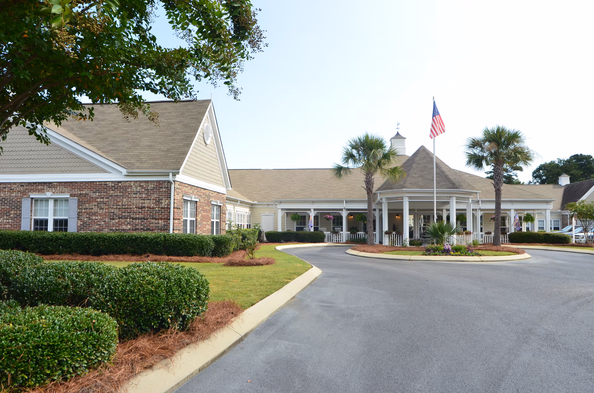 Front exterior of a single-story senior living facility with a circular driveway, porte-cochere, American flag, and palm trees.