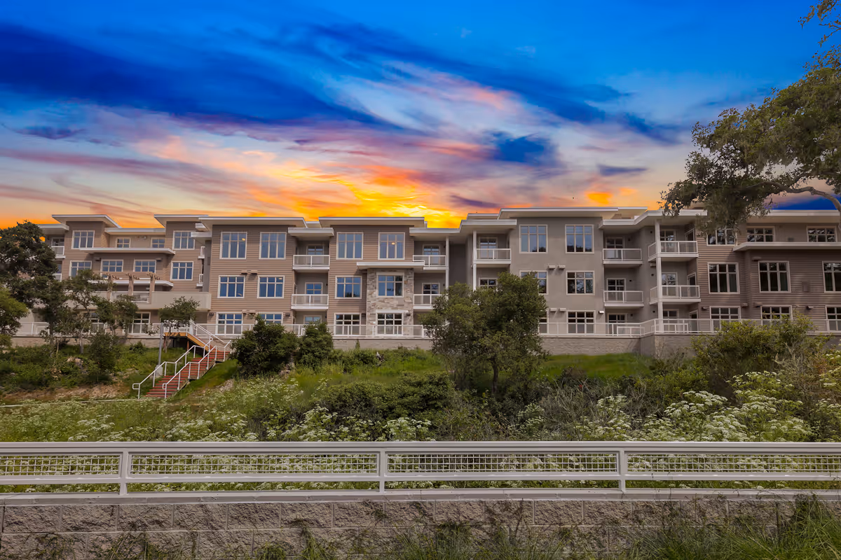 Exterior front view of a multi-story senior living building with landscaping under a colorful sunset sky.