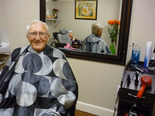 An elderly man with glasses sits in a barber chair wearing a black and gray patterned cape, smiling. Behind him is a large mirror reflecting his back and part of the room, including a vase with orange flowers and a framed picture on the wall. A hair dryer and other barber tools are visible on a counter to the right.