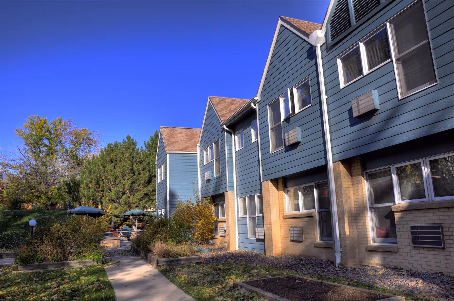 Exterior view of a two-story assisted living facility building with blue siding and brick lower walls, surrounded by trees and garden beds, with a clear blue sky overhead.