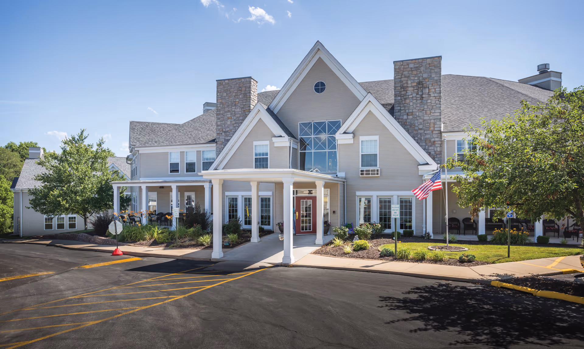 Front exterior of a large two-story senior living facility with a covered entrance, American flag, and landscaped grounds.