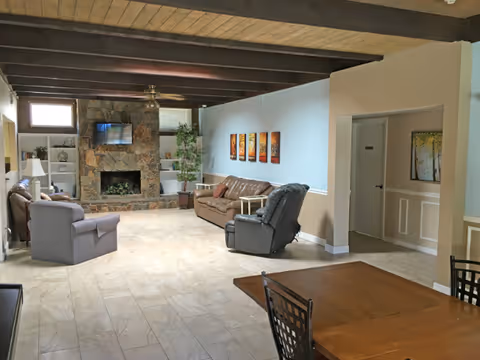 Spacious senior living common room with sofas and recliners facing a stone fireplace, exposed wood ceiling beams, and a dining table in the foreground.