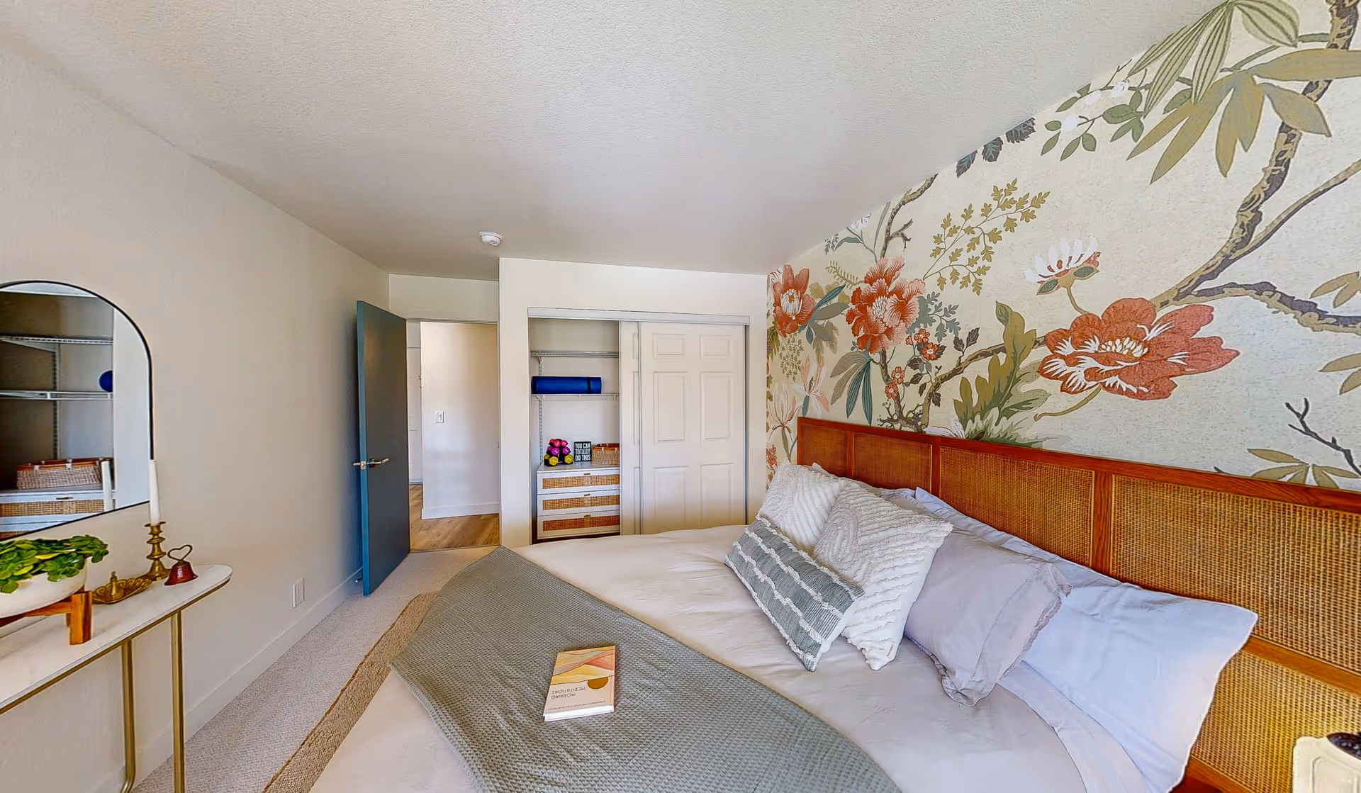 Bright bedroom with a floral accent wall, a made bed with pillows and a book, an open closet, and a small console table with a mirror.