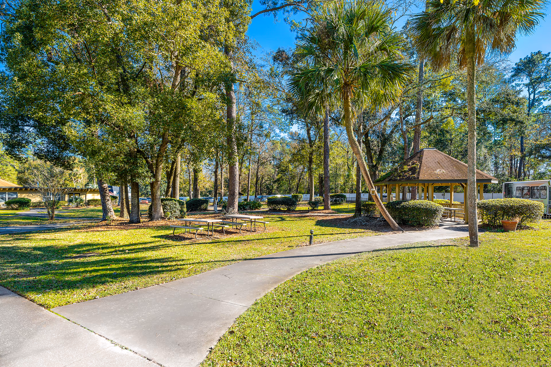 A sunny outdoor area at Riverwood Center featuring a paved walkway curving through a grassy lawn with trees and shrubs. There is a wooden gazebo with a shingled roof on the right side and picnic tables on the left side under the trees. A bus is partially visible in the background.