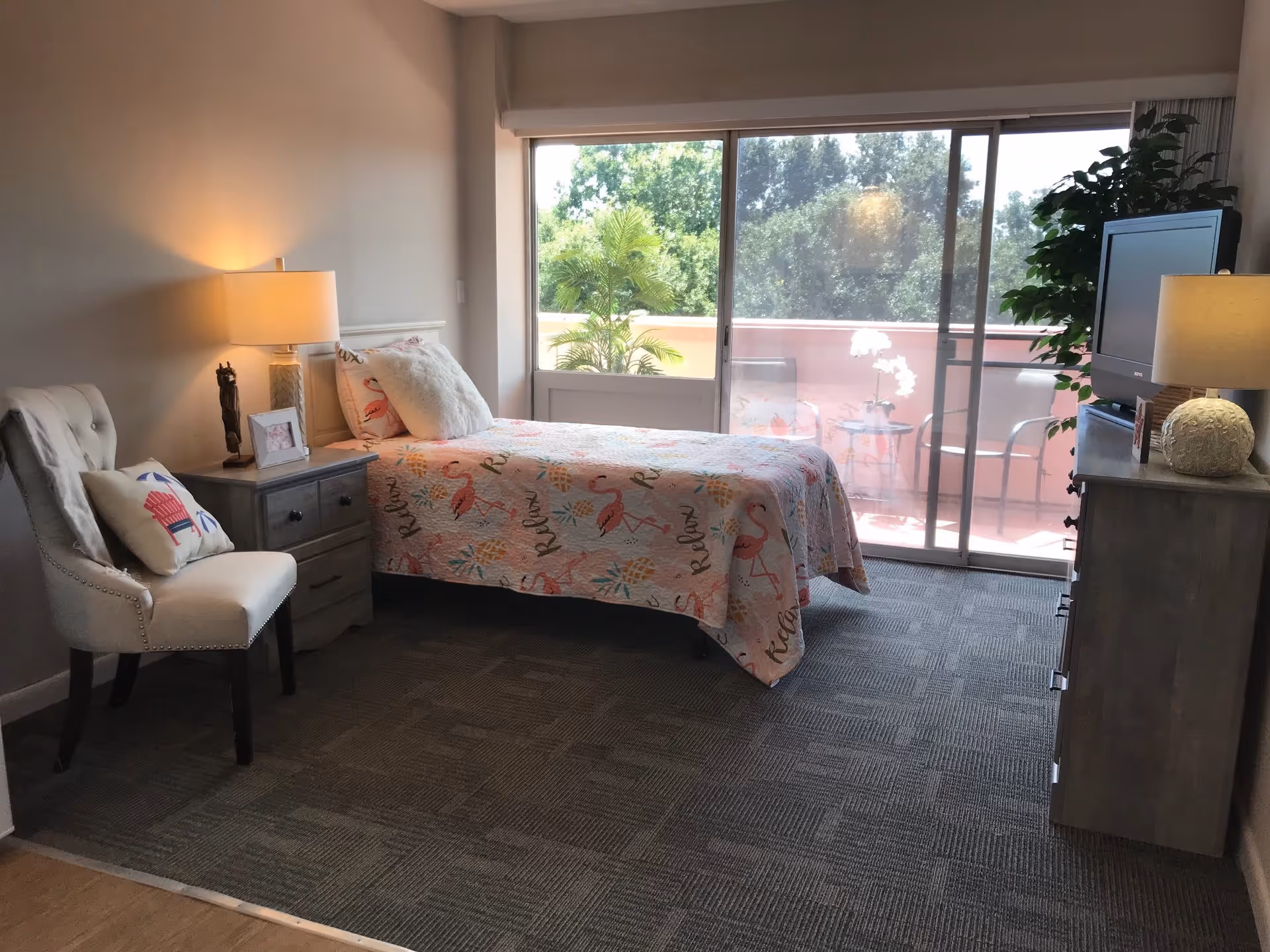 A bright and cozy bedroom with a single bed covered in a colorful flamingo-themed quilt. Next to the bed is a nightstand with a lamp, a small statue, and a framed photo. A cushioned chair with a decorative pillow is placed beside the nightstand. Across from the bed is a dresser with a TV and a lamp on top. Large sliding glass doors open to a balcony with outdoor chairs and a small table, with greenery visible outside.
