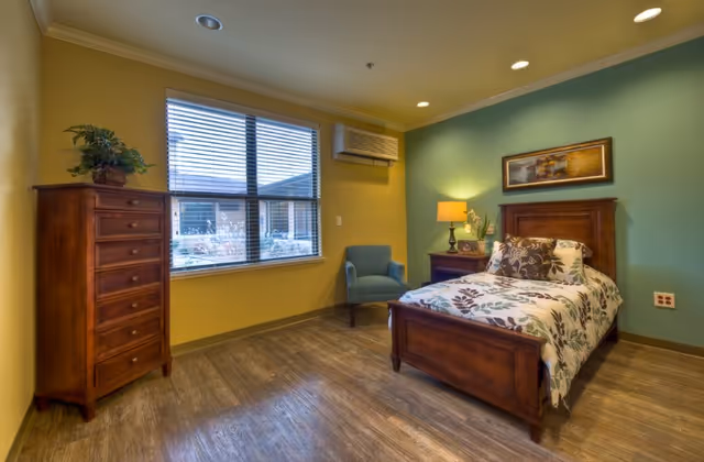 A cozy bedroom with a wooden bed featuring floral bedding, a wooden dresser with a plant on top, a blue armchair, a nightstand with a lamp and decorative items, a large window with blinds, and green and yellow painted walls.