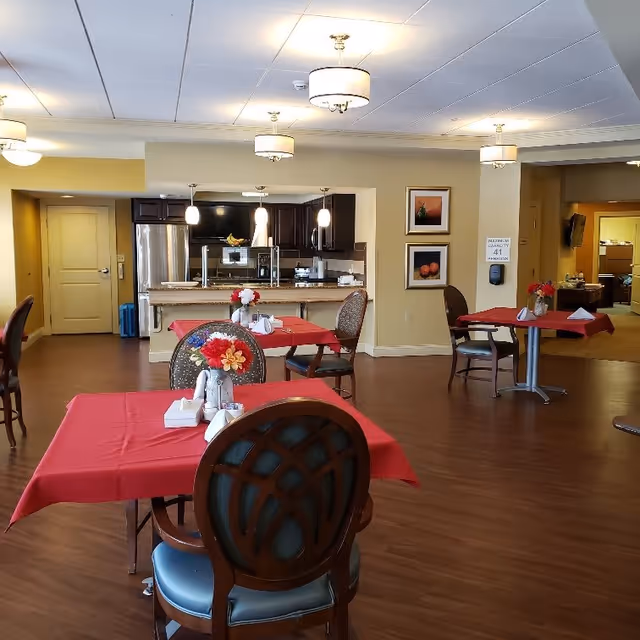A dining area in a senior living facility with several tables covered in red tablecloths, each set with napkins and small flower arrangements. The room has wooden flooring, pendant lights hanging over a kitchen counter with stainless steel appliances, and framed artwork on the walls.
