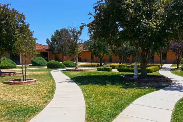 Curved concrete walkways surrounded by green grass, trees, and shrubs in an outdoor garden area of a senior living facility with a single-story building in the background under a clear blue sky.