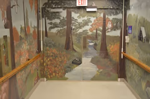 Hospital-style hallway with handrails leading to double doors painted with a woodland mural and an EXIT sign above.