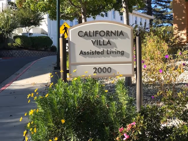 Outdoor view of a sign for California Villa Assisted Living with the number 2000, surrounded by green bushes and flowers, with a sidewalk and trees in the background.