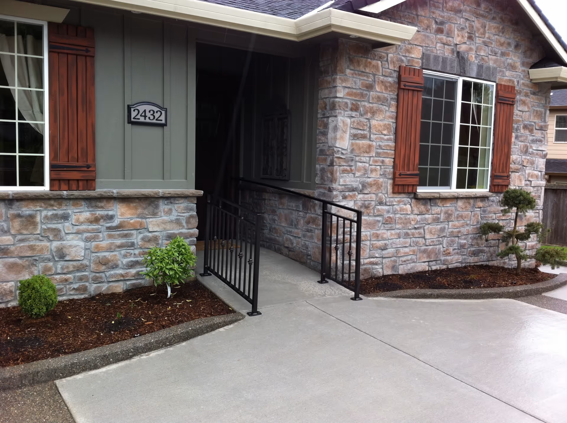 Exterior view of a stone and wood paneled building with a ramp entrance featuring black metal railings, two windows with wooden shutters, and small landscaped bushes along the concrete pathway.