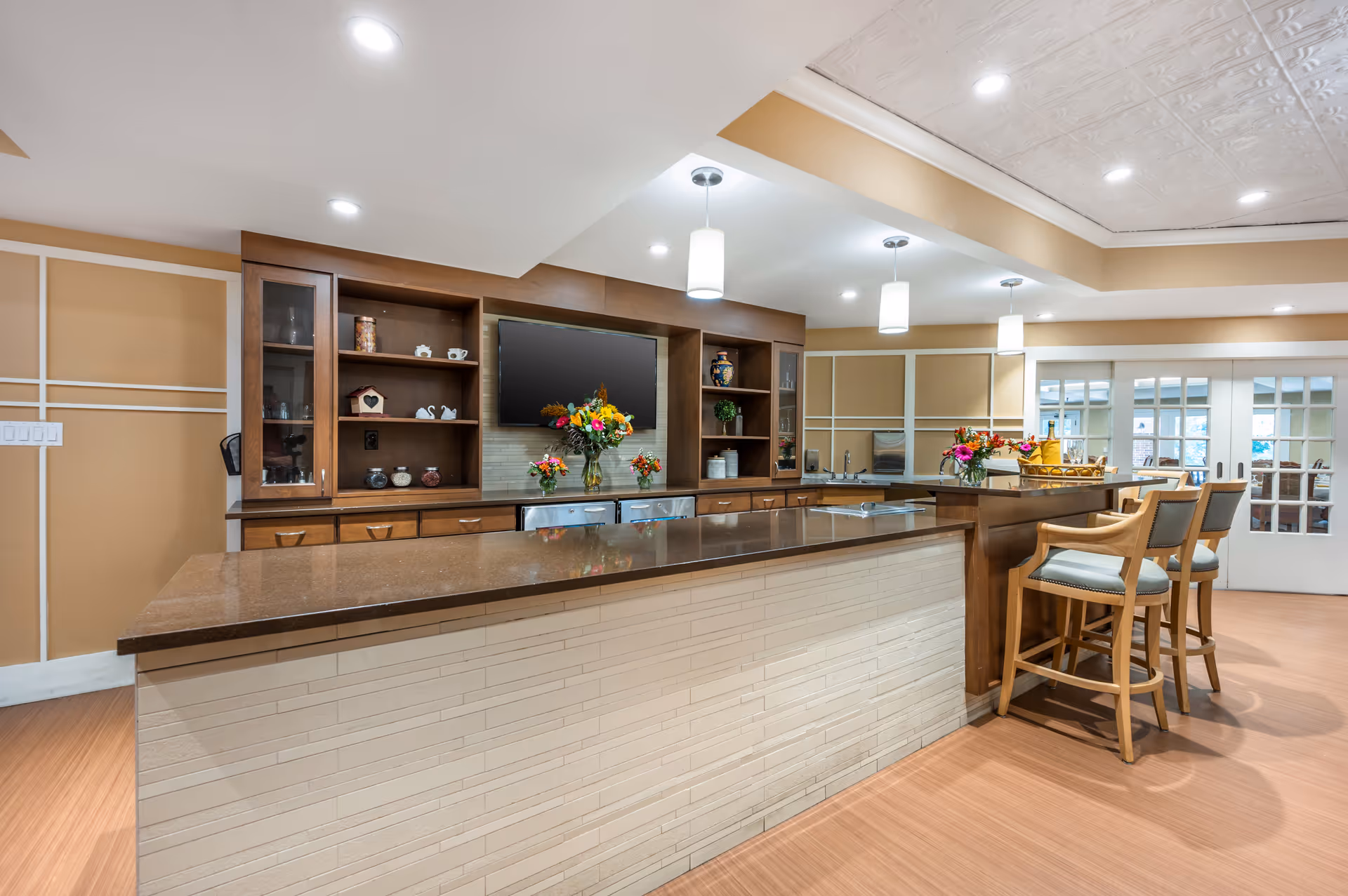 A modern kitchen area with a long brown countertop island and wooden cabinetry. The back wall features open shelves with decorative items and a mounted flat-screen TV. There are three pendant lights hanging from the ceiling and two wooden bar stools with light blue cushions at the counter. The room has beige walls with white trim and a patterned white ceiling. Sliding glass doors are visible in the background.