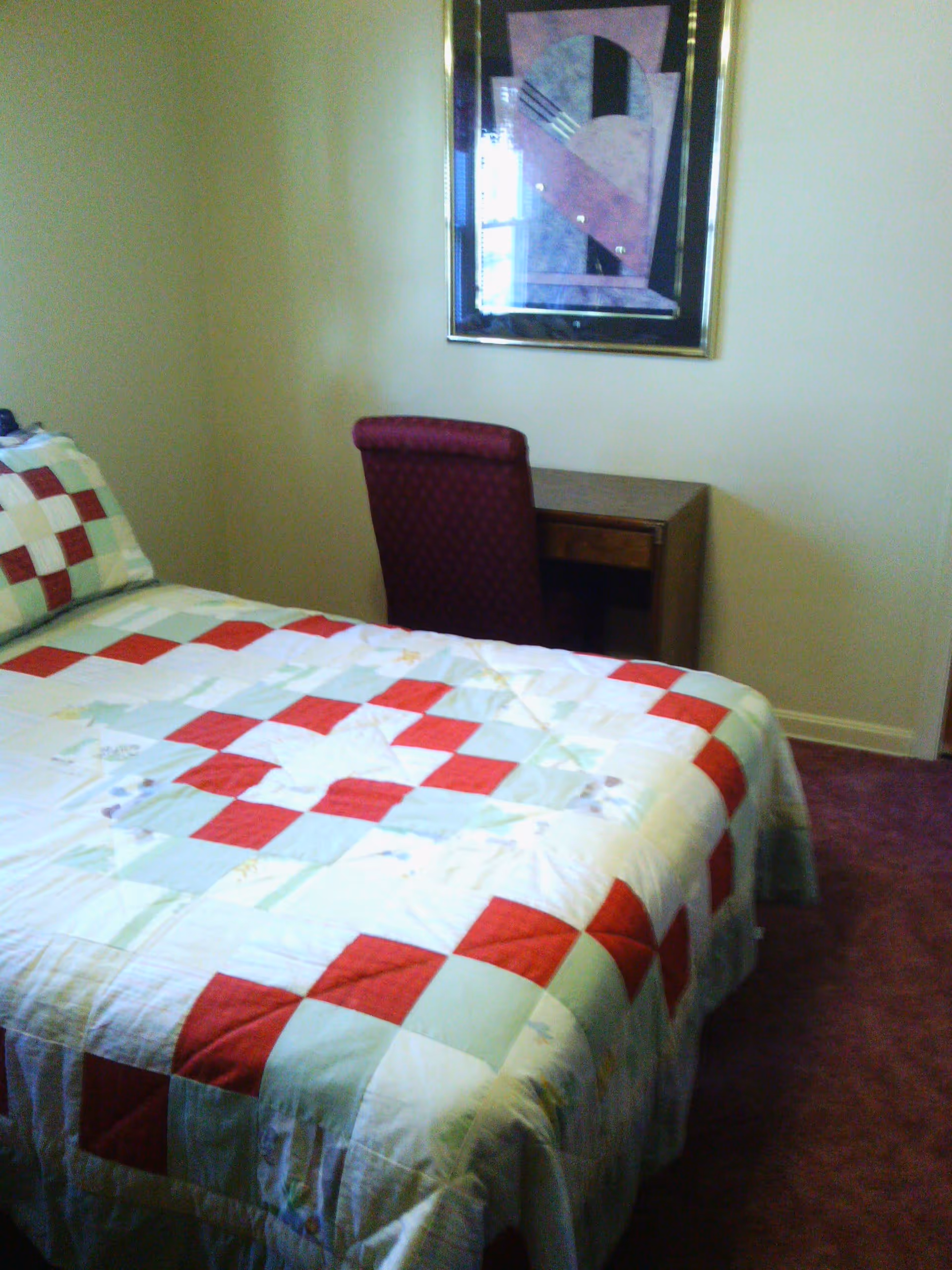A bedroom with a bed covered in a quilt with a red, white, and light blue checkered pattern. Next to the bed is a wooden desk with a maroon upholstered chair. A framed abstract painting hangs on the wall above the desk. The room has beige walls and a purple carpet.