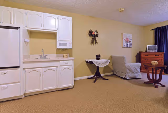 Interior view of a room featuring a small kitchenette with white cabinets, a sink, and a refrigerator on the left side. To the right, there is a small round table with a lace tablecloth and a decorative item on top, a cushioned armchair, a wooden dresser with a small TV on it, and a side table. The walls are painted beige, and there is a dark curtain covering a window on the far right.