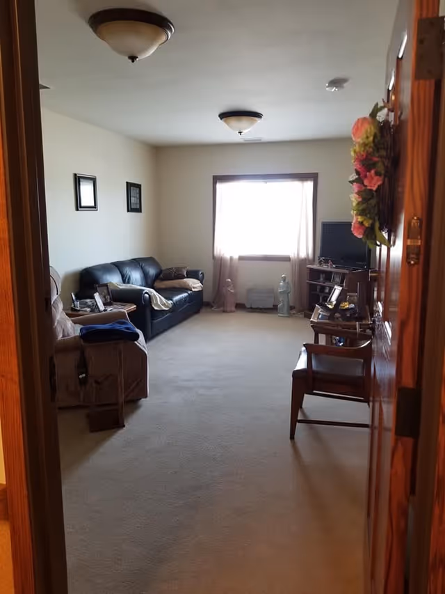 View into a living room with beige carpet and cream-colored walls. The room contains a black leather sofa with pillows, a beige recliner chair with a blue blanket, a wooden chair, a small wooden table with framed photos, a TV on a stand, and decorative statues near a window with sheer curtains. There is a floral wreath hanging on the wooden door in the foreground.