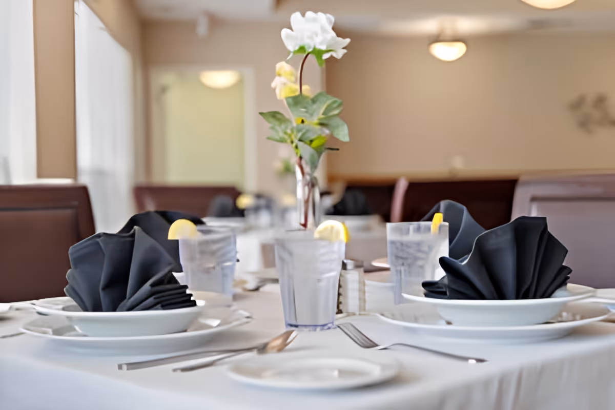 A dining table set with white plates, black folded napkins, glasses of water with lemon slices, silverware, and a vase with white and yellow flowers in a softly lit dining room.