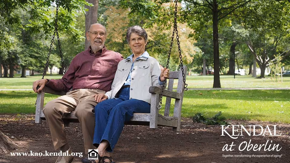 An elderly couple sitting together on a wooden swing in a park-like outdoor setting with green grass and trees in the background. The man is wearing a maroon shirt and beige pants, and the woman is wearing a light jacket and blue jeans. They are holding hands and smiling.