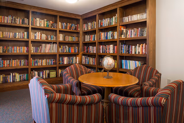 A cozy library room with wooden bookshelves filled with books lining the walls. In the center, there is a round wooden table with a globe on it, surrounded by four striped upholstered armchairs.