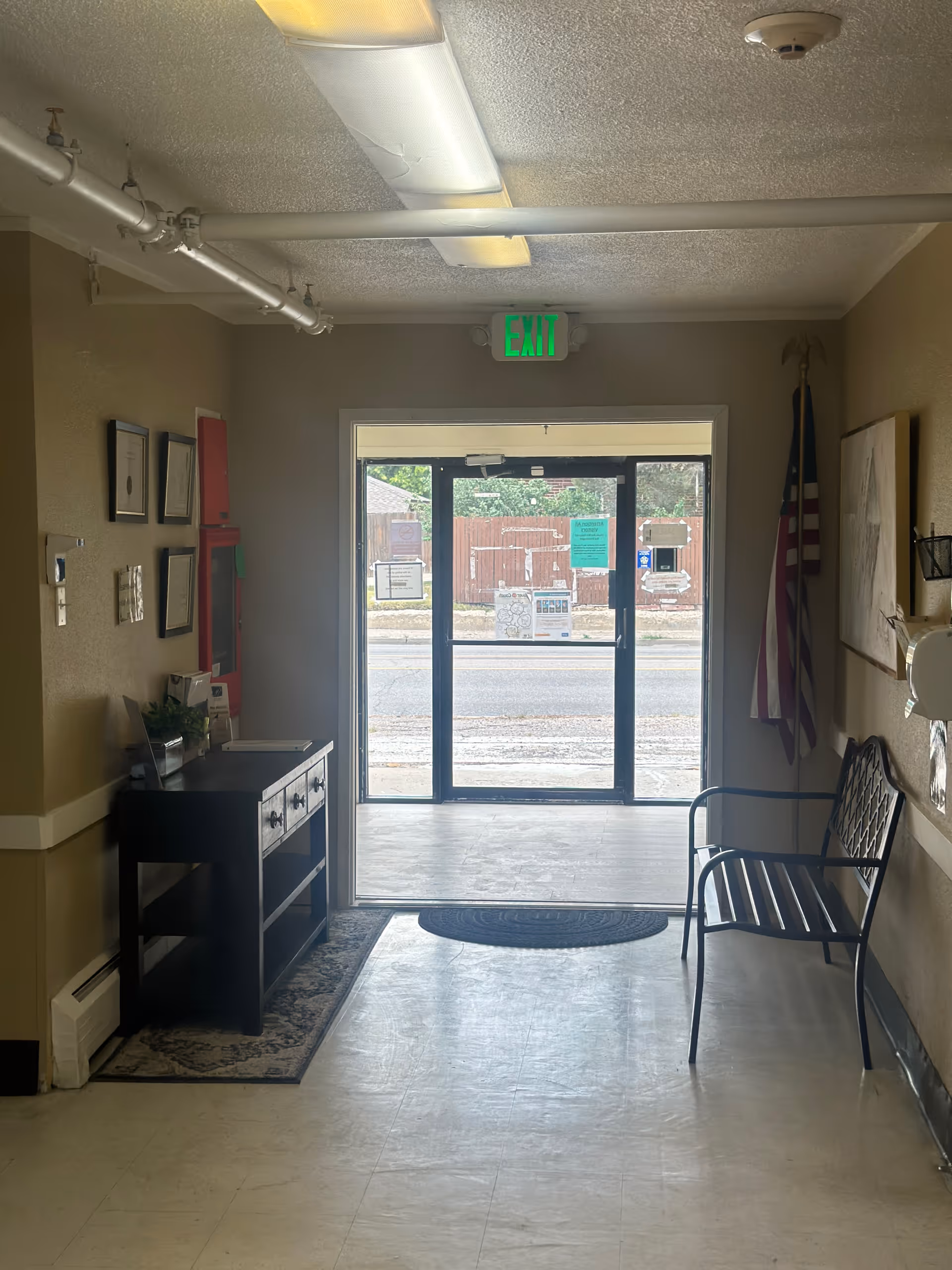 Interior hallway leading to glass exit doors with an 'EXIT' sign overhead, a bench on the right and a console table on the left.