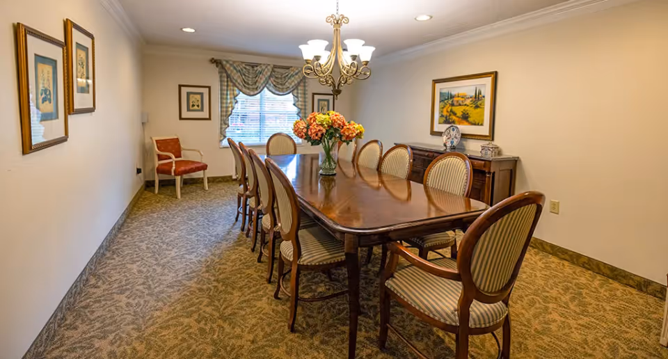 Formal dining room with a long wooden table, upholstered chairs, a chandelier and a floral centerpiece.