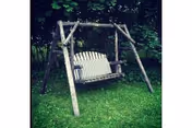 A wooden bench swing suspended from a rustic wooden frame on a grassy lawn with trees in the background.