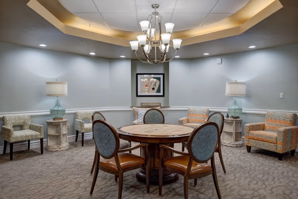 A cozy interior room with a round wooden table surrounded by six chairs with blue patterned backs and brown seats. The room has a beige carpet and light gray walls with white trim. There are two armchairs with orange and beige patterned upholstery and two additional chairs with light green patterned upholstery. Two small white side tables with glass lamps are placed beside the armchairs. A chandelier with multiple lights hangs from the ceiling, and a framed picture featuring playing cards is on the far wall.