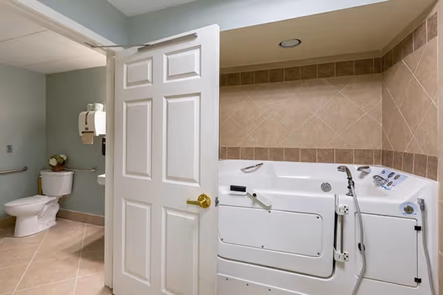 Bathroom with a white walk-in bathtub featuring safety handles and a handheld showerhead, beige tiled walls, and a white toilet with a grab bar next to it. The door to the bathroom is partially open.