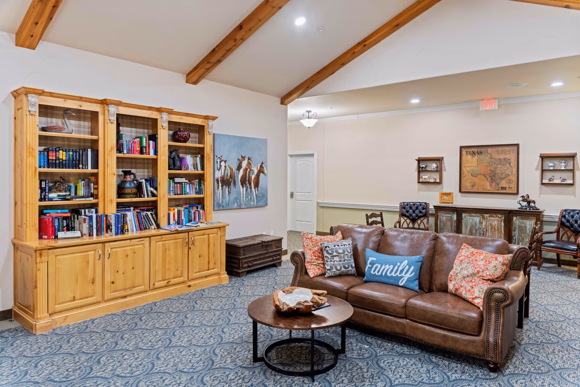 A cozy senior living common area featuring a brown leather sofa with decorative pillows, including one that says 'Family'. In front of the sofa is a round wooden coffee table with a decorative bowl. To the left, there is a large wooden bookshelf filled with books and decorative items. The room has a patterned carpet, exposed wooden ceiling beams, and soft lighting. On the far wall, there is a painting of horses and a framed map of Texas with shelves holding small decorative items.